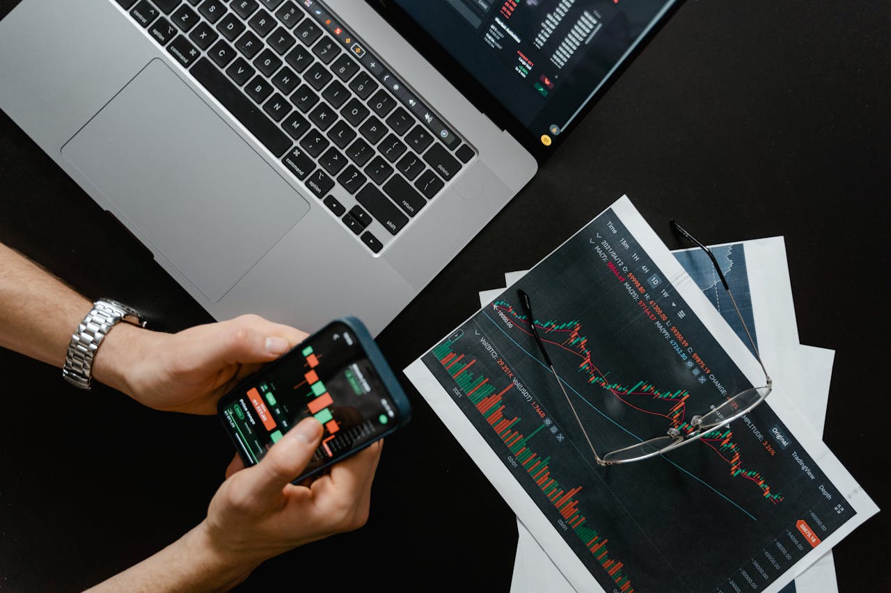 Hands using smartphone beside laptop with stock charts, showcasing digital trading.